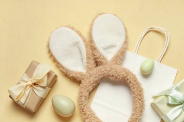 Tavşan kulaklı alışveriş çantası, hediye kutuları ve sarı arka planda Paskalya yumurtaları.