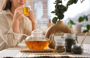 Mutfakta çay içen kadına karşı yemek masasında okaliptüs dalları ve çaydanlığı olan bir vazo.