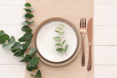 Tabaklar, çatal bıçak takımları ve okaliptüs dallarıyla beyaz ahşap arka planda şık bir masa.