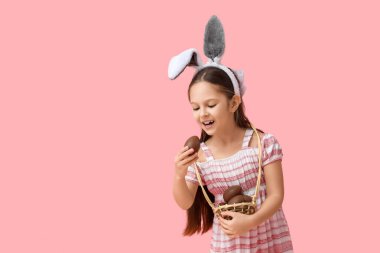 Tavşan kulaklı şirin bir kız elinde sepetle pembe arka planda çikolata yumurtaları tutuyor. Paskalya kutlaması