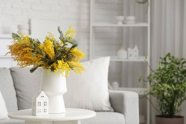Mimoza çiçekli vazo ve oturma odasındaki masada duran ev figürü.