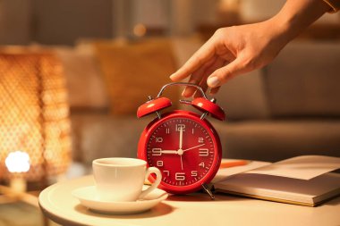 Kadın, akşam vakti odada kitap ve bir fincan kahveyle masadaki çalar saati kapatıyor.