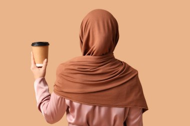 Kahverengi arka planda, kağıt bardak sıcak kahveyle tesettürlü Müslüman kadın.