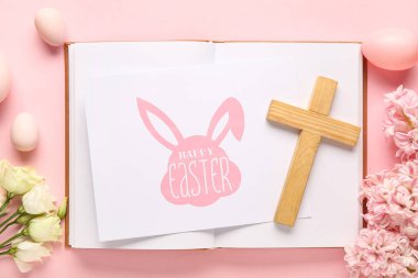 Kitap, yumurta, çiçek ve pembe arka planda Mutlu Paskalyalar tebrik kartı olan tahta haç.