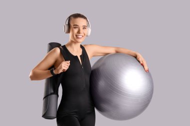 Kulaklıklı, spor kulaklıklı, gri arka planda yoga minderi olan genç bir kadın.