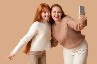 Genç kızıl kadın ve tatlı kızı bej arka planda selfie çekiyorlar.