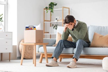 Evdeki kanepede oturan bir kutu kişisel eşyası olan kovulmuş genç adam.