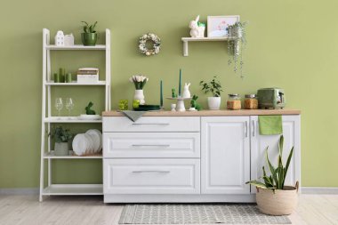 Mutfağın içinde raflar, Paskalya çelengi, çiçekler ve çatal bıçaklar yeşil duvarın yanında.