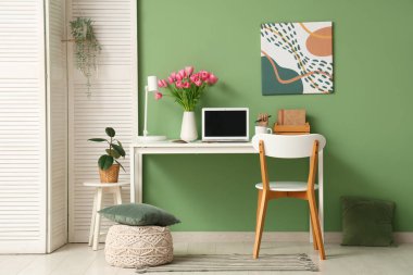 Modern dizüstü bilgisayarı, kitapları ve yeşil duvarın yanındaki güzel lale çiçekleriyle süslü çalışma alanı.