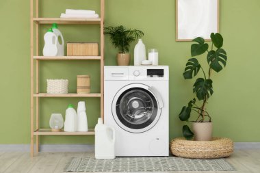 Çamaşır odasındaki yeşil duvarın yanında raf ünitesi, ev bitkileri ve deterjanları olan modern çamaşır makinesi.