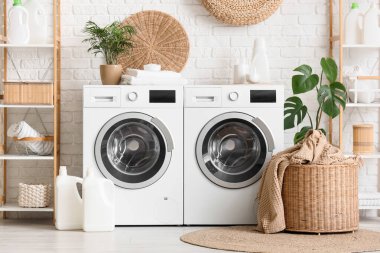 Çamaşır odasındaki beyaz tuğla duvarın yanında çamaşır makinesi, sepet ve deterjan.