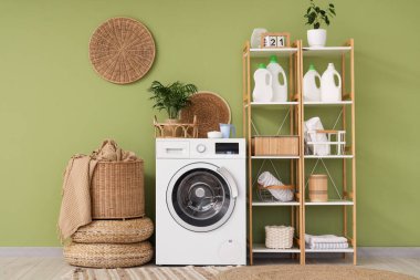 Çamaşır odasındaki yeşil duvarın yanında raflar, sepet ve deterjanlar olan modern çamaşır makinesi.