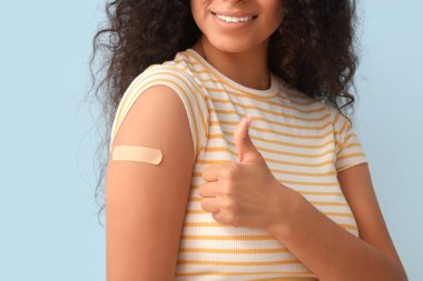 Tıbbi yaması olan genç bir kadın mavi arka plandaki aşıdan sonra baş parmağını kaldırmış.