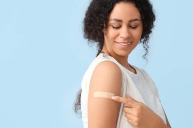 Genç Afro-Amerikalı kadın mavi arka plandaki aşıdan sonra tıbbi yamaya işaret ediyor.