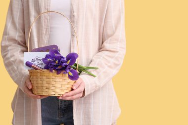 Sarı arkaplanda anneler günü için çiçek, kartpostal ve kozmetik taşıyan sepet taşıyan genç bir kadın.