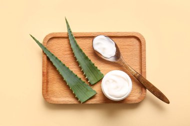 Kavanoz ve ahşap kaşık dolusu kozmetik krem. Bej arka planda aloe vera yaprakları var.