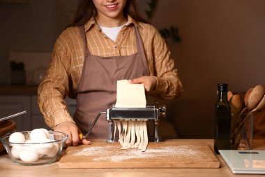 Genç bir kadın karanlık mutfakta makineyle makarna yapıyor.