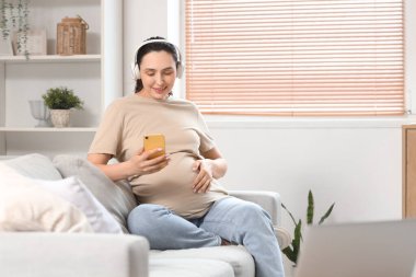 Kulaklıklı genç hamile kadın evdeki koltukta cep telefonu kullanıyor.
