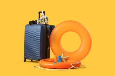 Şişirilebilir yüzüklü bavul, can simidi, güneş kremi ve sarı arka planda plaj aksesuarları. Tatil kavramı