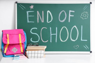 Sırt çantalı, kitaplı ve açık renkli duvarın yanındaki tahtada Okulun Sonu yazılı masa