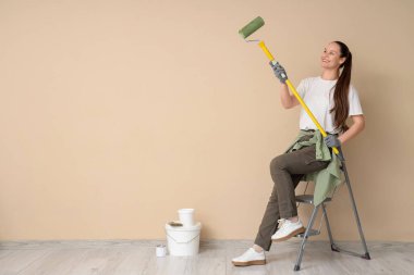 Boya silindirli genç bir kadın bej duvarın yanındaki merdivende oturuyor.