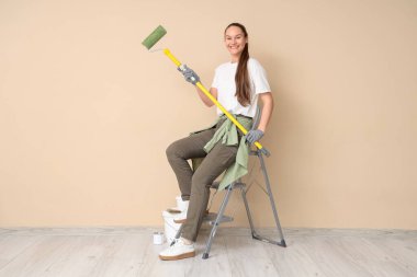 Boya silindirli genç bir kadın bej duvarın yanındaki merdivende oturuyor.