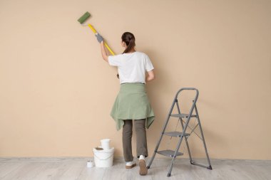 Tekerlekli sandalyesi ve merdiveni olan, bej renkli duvarı boyayan genç bir kadın.