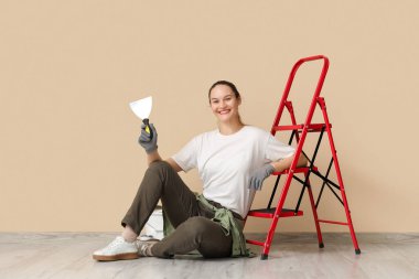 Bej rengi duvarın yanında oturan elinde oyuncak bıçak ve merdiven olan genç bir kadın.