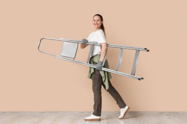 Genç bir kadın bej duvarın yanında merdiven taşıyor.