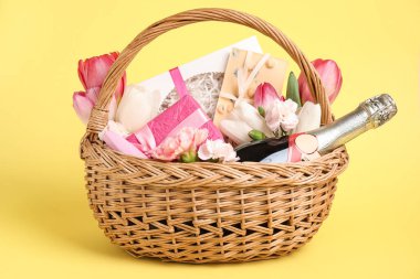 Şampanya şişesi, hediye kutuları, çikolata ve sarı arka planda bahar çiçekleri olan hasır sepet.
