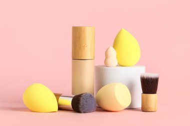 Pembe arka planda fırçalı makyaj süngeri ve bir şişe fondöten.