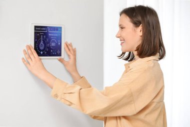 Genç, mutlu bir kadın odasındaki duvarda akıllı ev güvenlik kontrol paneli kullanıyor.