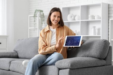 Genç mutlu kadın, odasındaki kanepede akıllı ev güvenlik kontrol paneliyle tablet tutuyor.