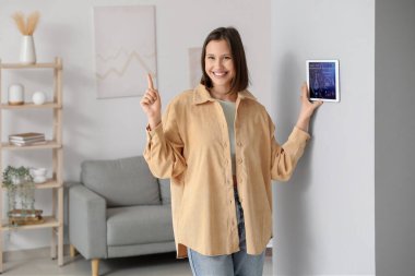 Genç mutlu kadın duvarda akıllı ev güvenlik kontrol paneli kullanıyor ve odadaki bir şeyi işaret ediyor.
