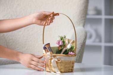 Hasır sepet kozmetik ürünleri, hediye kutuları ve evdeki masada çiçekleri olan genç bir kadın.