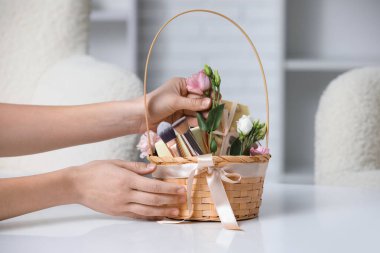 Hasır sepet kozmetik ürünleri, hediye kutuları ve evdeki masada çiçekleri olan kadın elleri, yakın plan.