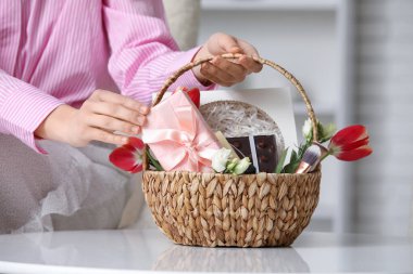 Kozmetik ürünleri, hediye kutusu, çikolata ve çiçeklerle dolu sepeti evdeki masada tutan genç bir kadın.