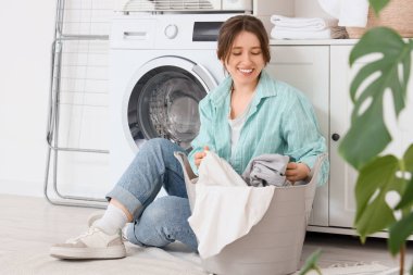 Çamaşır odasında çamaşır sepetinin yanında oturan genç bir kadın.