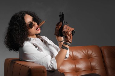Siyah arka planda kanepede otururken elinde silahla puro içen seksi Afro-Amerikalı kadın.
