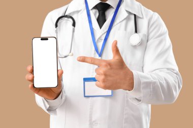 Erkek doktor, bej arka plandaki boş cep telefonunu işaret ediyor, yakın plan.