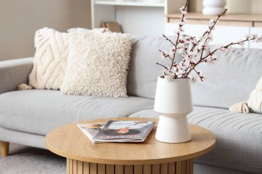 Oturma odasındaki kanepenin yanındaki sehpanın üzerinde dergiler olan çiçek açan bir vazo.