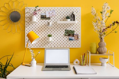 Boş dizüstü bilgisayarlı işyeri, çiçek açan dallar ve sarı duvardaki tahta ofis.