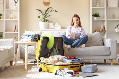 Evdeki kanepede toplanacak eşyaların listesini hazırlayan valizli kadın turist.