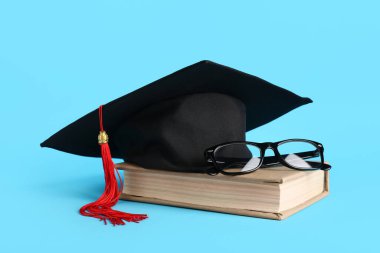 Mezuniyet şapkası, kitap ve gözlükleri mavi arka planda.