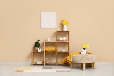 Oturma odasındaki bej duvarın yanında puf ve narsis çiçekli raf ünitesi.