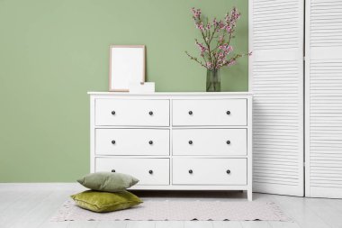 Üzerinde fotoğraf çerçevesi, giyinme ekranı ve yeşil duvarın yakınındaki pembe çiçek açan vazolardan oluşan bir elbise dolabı.