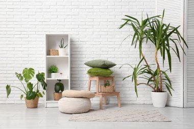 Oturma odasındaki beyaz tuğla duvarın yanında merdiven, yastık ve farklı ev bitkileri olan raf ünitesi.