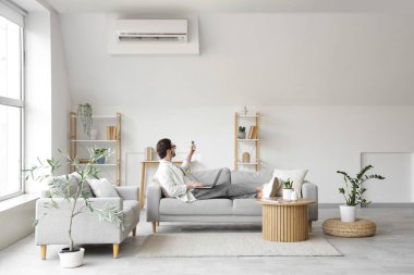 Dizüstü bilgisayarı olan genç adam oturma odasında klimayı açıyor.