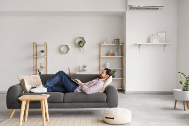 Genç adam kanepede dizüstü bilgisayar kullanıyor ve oturma odasında klima var.