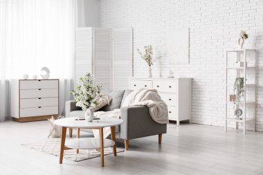 Şifoniyerli kanepe, sehpa ve oturma odasındaki beyaz tuğla duvarın yanında çiçek açan dalların olduğu bir vazo.